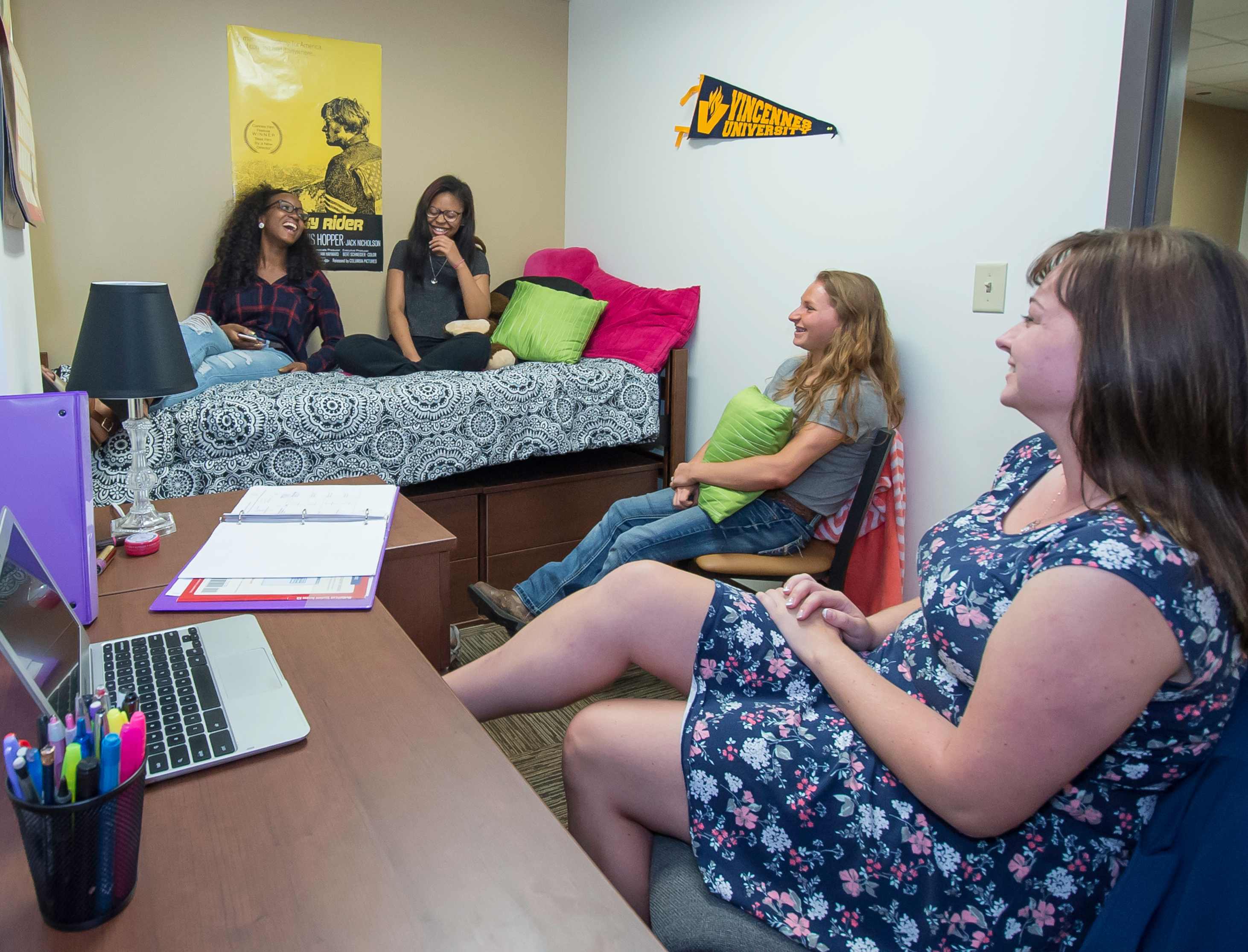 A diverse group of female students sit on beds and chairs laughing in VU residence hall room 