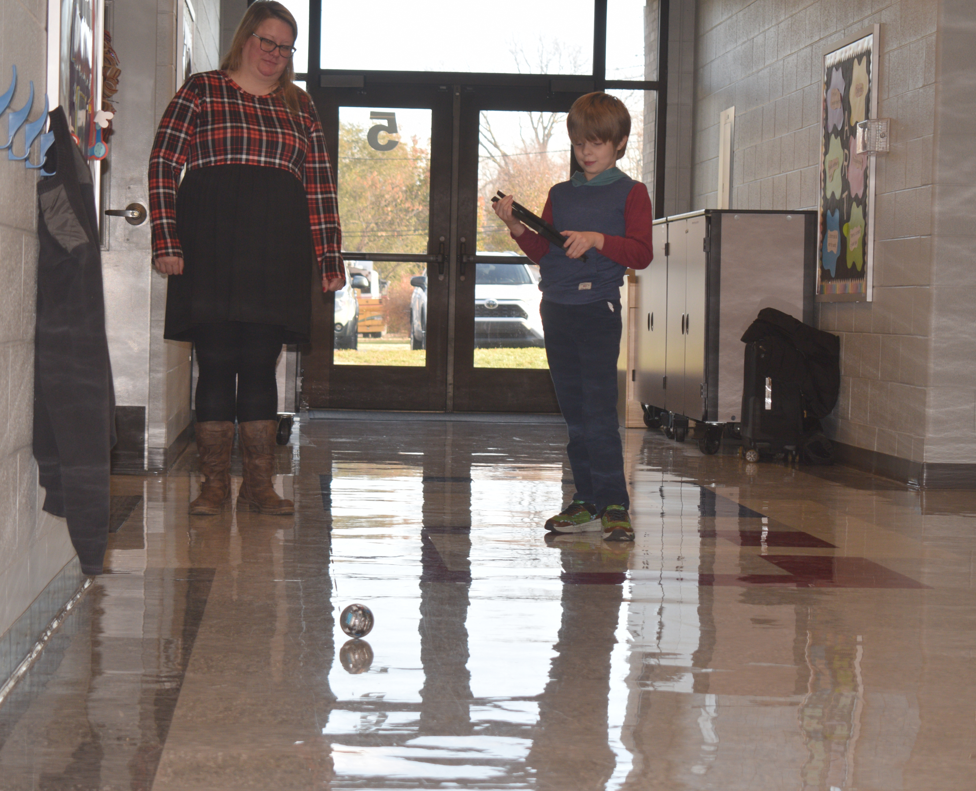 A male student holding a laptop interacts with a Sphero BOLT Coding Robot in a hallway at Tecumseh-Harrison School.