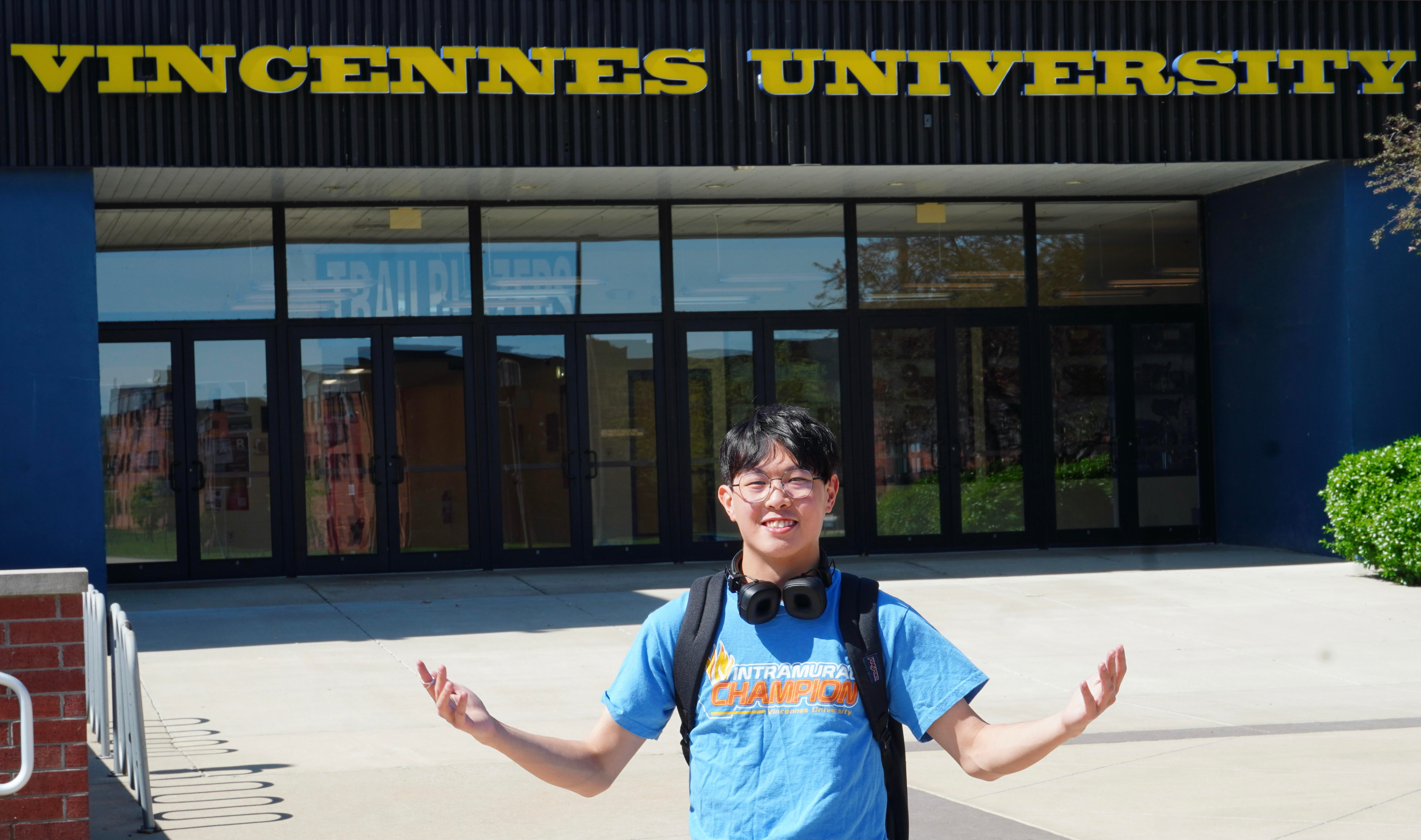 Samuel Lo stands with his arms extended in front of an entrance to the P.E. Complex. Vincennes University is displaced over the doors.er 