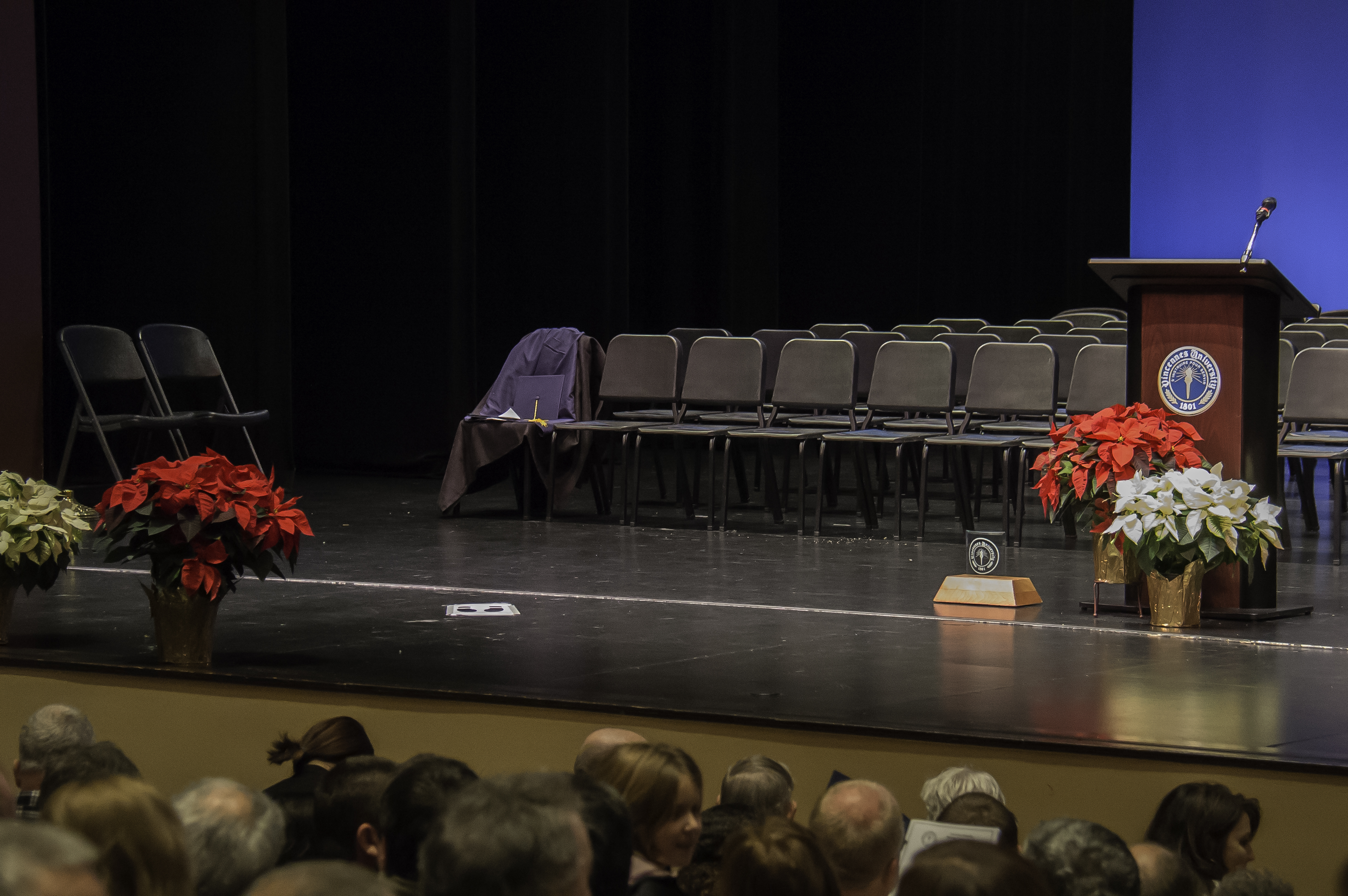 Chairs, poinsettias, and a podium on the Red Skelton Performing Arts Center stage. A cap and gown are on one of the chairs. Male and female guests are sitting in the audience in front of the stage.